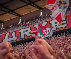 Fans beim Singen in der Südkurve. Im Hintergrund die Zaunfahne der Südkurve.