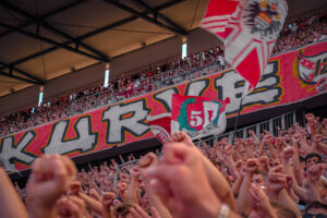 Fans beim Singen in der Südkurve. Im Hintergrund die Zaunfahne der Südkurve.